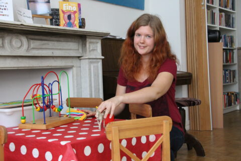 Punatukkainen hymyilevä nuori nainen lasten pöydän äärellä leluja kädessään.