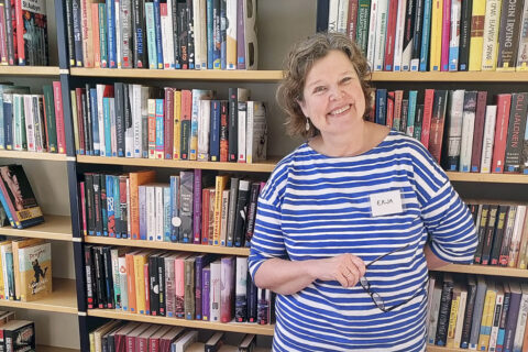 Hymyilevä sinivalkoraitapaitainen nainen kirjahyllyn edessä.