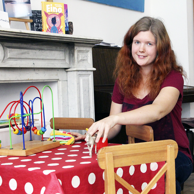 Punatukkainen hymyilevä nuori nainen lasten pöydän äärellä leluja kädessään.