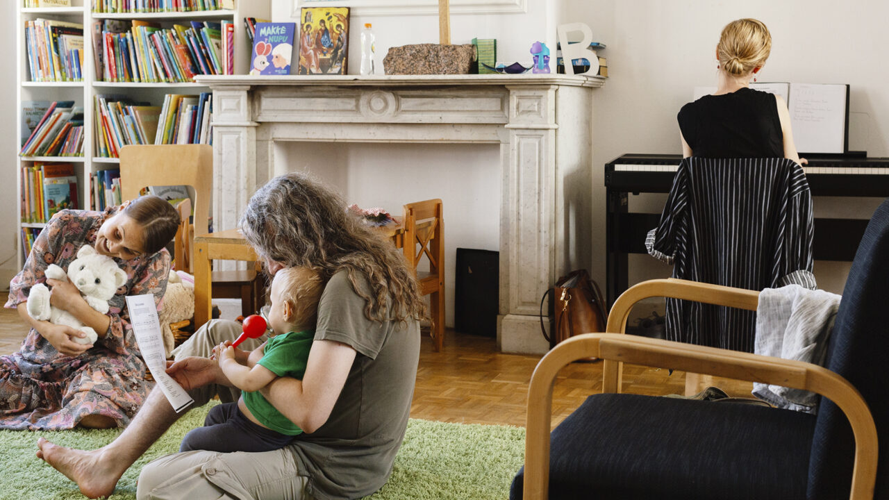 Kaksi äitiä lapsi sylissään istuu lattialla ja taustalla nainen soittaa pianoa.